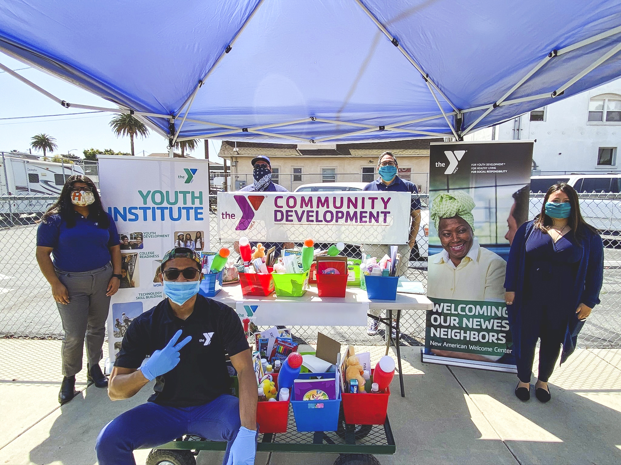 A diverse group of people wearing masks and gloves standing and sitting around some nright colored bins full of children's toys. There are 3 signs in the image, one says 