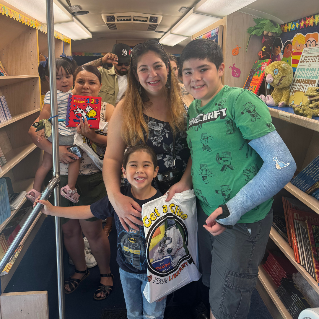 Familys are aboard a bus with shelves lined with books. They are smiling and holdding bags and books as they exit the vehicle